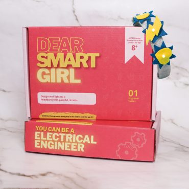 Dear Smart Girl Electrical Engineer STEM Kit standing on top of another of the same kit. Light-up headpiece hanging off of the top right corner of standing box.
