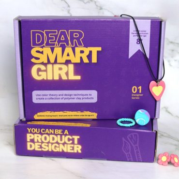 Dear Smart Girl Product Designer STEM Kit box standing on another of the same kit. Polymer clay jewelry around and on top of box.